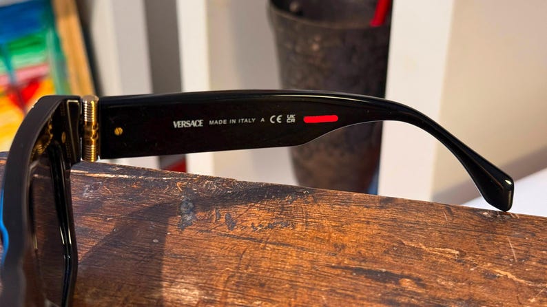 May include: Black Versace sunglasses with gold accents. The text "VERSACE MADE IN ITALY" is printed on the arm. A red stripe is visible on the arm. The sunglasses are resting on a wooden surface.