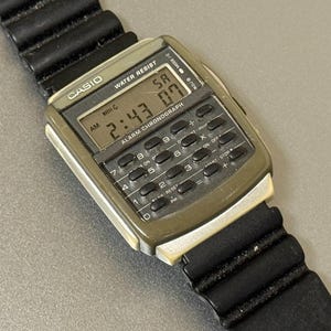 May include: A vintage Casio calculator watch with a black rubber strap. The watch face displays the time as 2:43 and the date as 07. The watch has a calculator keypad and the Casio logo is visible.