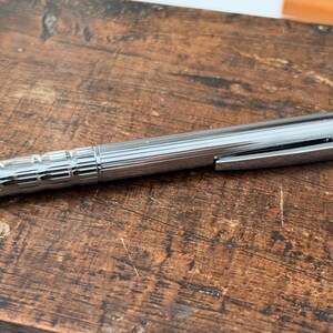 Op de afbeelding: Een zilverkleurige metalen balpen met een textuur gripgedeelte met horizontale groeven, een gladde houder met verticale strepen, een zakclip en een opvallend rood ringaccent bij het drukmechanisme aan de bovenzijde.