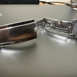 May include: A silver watch band with a clasp that is open. The band is made of metal links.