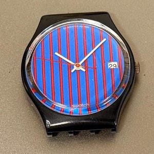 Puede incluir: Un reloj Swatch negro con esfera a rayas azules y rojas. El reloj tiene una pantalla de fecha que muestra el número 29. Las manecillas del reloj son blancas con un centro rojo. El texto "Swatch AD 1986" indica que es un modelo vintage.