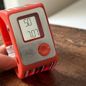 Op de afbeelding: Een rood-wit gestreept digitaal polshorloge met een zilveren wijzerplaat en een rode knop. Het horloge geeft de tijd 50:07 aan.