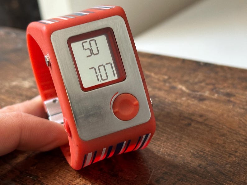 Op de afbeelding: Een rood-wit gestreept digitaal polshorloge met een zilveren wijzerplaat en een rode knop. Het horloge geeft de tijd 50:07 aan.
