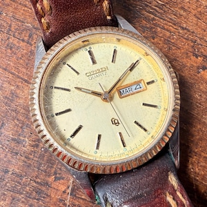 Può includere: Un orologio da polso Citizen Quartz color oro con cinturino in pelle marrone. Il quadrante dell'orologio è bianco con segni neri e una finestra della data che indica "MAR 4".