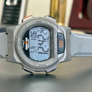 K&ouml;nnte beinhalten: Eine Timex Ironman Digitaluhr mit grauem Armband. Das Zifferblatt zeigt die Uhrzeit und andere Daten an. Die Uhr verf&uuml;gt &uuml;ber Tasten f&uuml;r verschiedene Funktionen, darunter Einstellen, Abrufen, Modus, Start und Split. Die Uhr ist f&uuml;r den Triathlon-Einsatz konzipiert.