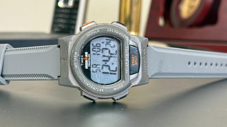 K&ouml;nnte beinhalten: Eine Timex Ironman Digitaluhr mit grauem Armband. Das Zifferblatt zeigt die Uhrzeit und andere Daten an. Die Uhr verf&uuml;gt &uuml;ber Tasten f&uuml;r verschiedene Funktionen, darunter Einstellen, Abrufen, Modus, Start und Split. Die Uhr ist f&uuml;r den Triathlon-Einsatz konzipiert.