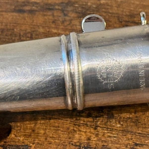 Pode incluir: Uma boquilha de saxofone de metal prateado com uma faixa texturizada, uma pequena alavanca saliente e um logótipo gravado que diz Henri Selmer Paris com o texto Made in France visível na lateral do componente cilíndrico do instrumento.