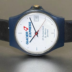 May include: A blue Glacier Express watch with a white face, black hands, and a red second hand. The watch face displays the date and the Glacier Express logo. The watch has a black leather band.