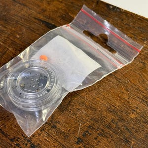 May include: A small clear plastic container with a black lid containing a small amount of black glitter. The container is in a clear plastic bag with a red zipper.