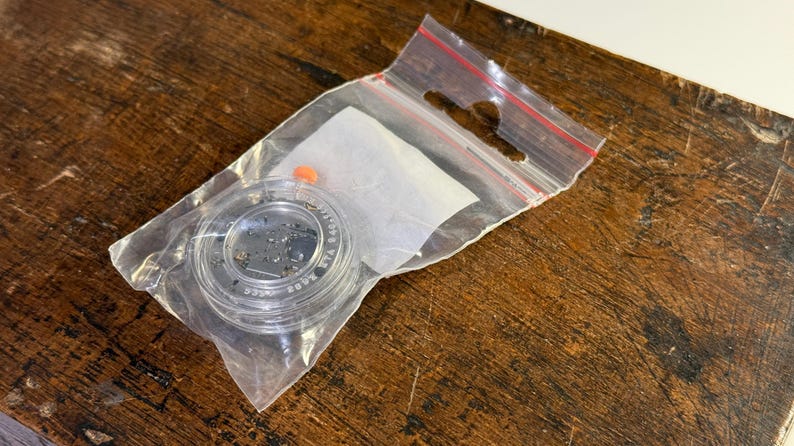 May include: A small clear plastic container with a black lid containing a small amount of black glitter. The container is in a clear plastic bag with a red zipper.