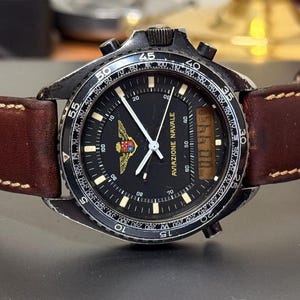Könnte beinhalten: Eine schwarze analog-digitale Armbanduhr mit einem braunen Lederarmband. Das Zifferblatt der Uhr hat einen schwarzen Hintergrund mit weißen Zeigern und Markierungen. Die Uhr hat eine drehbare Lünette mit weißen Markierungen. Das Zifferblatt der Uhr hat auch ein kleines digitales Display mit dem Text "AVIAZIONE NAVALE".