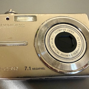 May include: A silver Olympus FE-230 digital camera featuring a 7.1 megapixel sensor and an Olympus lens with 3x optical zoom, 6.3-18.9mm, 1:3.1-5.9. The compact device includes a built-in flash, a small red indicator light, and a horizontal silver bar.