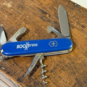 Può includere: Un coltellino svizzero blu con un logo bianco che dice "BOOKxpress". Il coltellino ha una varietà di strumenti, tra cui una lama, un cavatappi, un apribottiglie e un cacciavite.