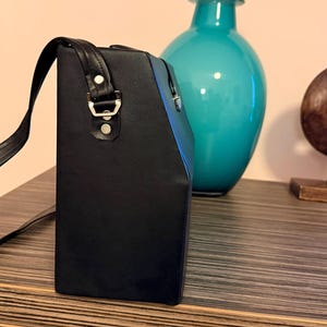 Puede incluir: Un bolso negro rectangular con una correa para el hombro. El bolso tiene herrajes de color plateado y está hecho de un material liso. Un jarrón turquesa y una escultura de madera están en el fondo.
