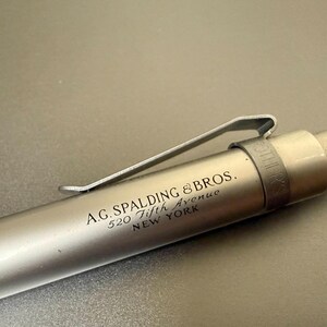Könnte beinhalten: Ein silberfarbener Stift mit einem Metallclip. Auf dem Stift steht "A.G. SPALDING & BROS. 520 Fifth Avenue NEW YORK". Der Stift liegt auf einer grauen Oberfläche.