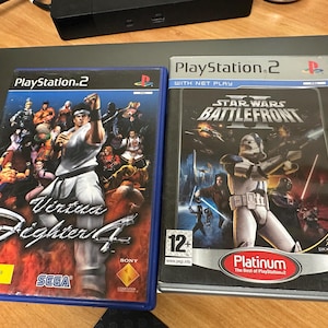 Puede incluir: Dos cajas de juegos de video para PlayStation 2. La caja de la izquierda es azul y presenta el juego Virtua Fighter 4. La caja de la derecha es negra y presenta el juego Star Wars Battlefront II. Ambas cajas tienen una clasificación de 12+.