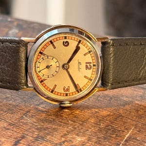 Puede incluir: Un reloj de pulsera vintage dorado con una correa de cuero negro. La esfera del reloj es de color crema con números y manecillas negras, y un pequeño dial de segundos. El reloj dice "Mido".