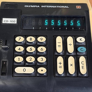 May include: A vintage Olympia International CD 100 desktop calculator with a beige casing and a black control panel. The device features a digital display showing the number 55555, a numeric keypad, and function keys for arithmetic operations.
