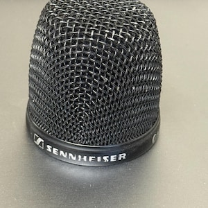 May include: A black microphone head with a mesh grille. The base of the microphone head has the brand name "SENNHEISER" printed in white. The microphone head is cylindrical and sits on a gray surface.