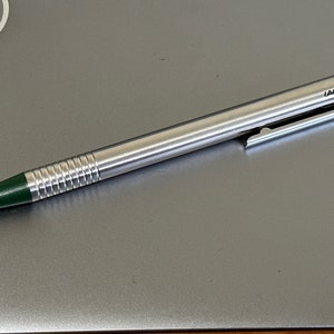 Puede incluir: Un bol&iacute;grafo plateado con una punta verde y un clip verde. El bol&iacute;grafo tiene un cuerpo acanalado y la palabra "LAMY" est&aacute; impresa en el clip.