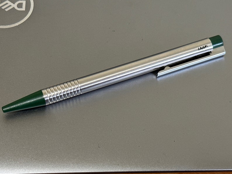 Puede incluir: Un bol&iacute;grafo plateado con una punta verde y un clip verde. El bol&iacute;grafo tiene un cuerpo acanalado y la palabra "LAMY" est&aacute; impresa en el clip.