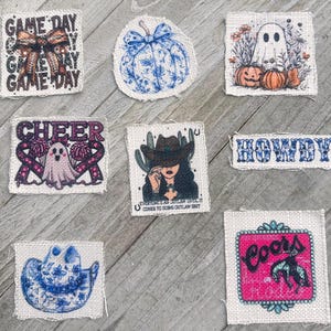 May include: A collection of eight fabric patches with various designs, including a pumpkin, a ghost, a cowboy hat, a cheerleading motif, a cowboy boot, and a Coors beer logo. The patches are all on a light brown wooden surface.