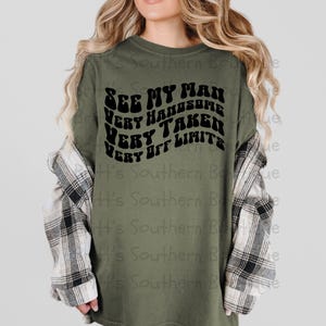 May include: A green long-sleeved shirt with a black graphic that reads "See My Man Very Handsome Very Taken Very Off Limits" in a retro font.