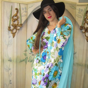 May include: A long, white maxi dress with a floral print in shades of blue, purple, green, and yellow. The dress has a V-neckline and long sleeves. The model is wearing a black hat and a turquoise scarf.