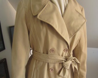 Könnte beinhalten: Ein beigefarbener, doppeltbreit gefütterter Trenchcoat mit passendem Gürtel, der in der Taille gebunden ist. Der Mantel hat einen Reverskragen, Knopfverschlüsse und ein klassisches Design. Der Mantel ist aus einem glatten Stoff gefertigt und hat eine taillierte Passform.