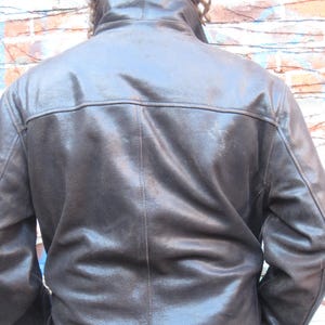 May include: A dark brown leather jacket with a high collar and a glossy finish. The jacket has a classic design with visible stitching and a tailored fit. The image shows the back of the jacket, highlighting its structured shoulders and sleek silhouette.