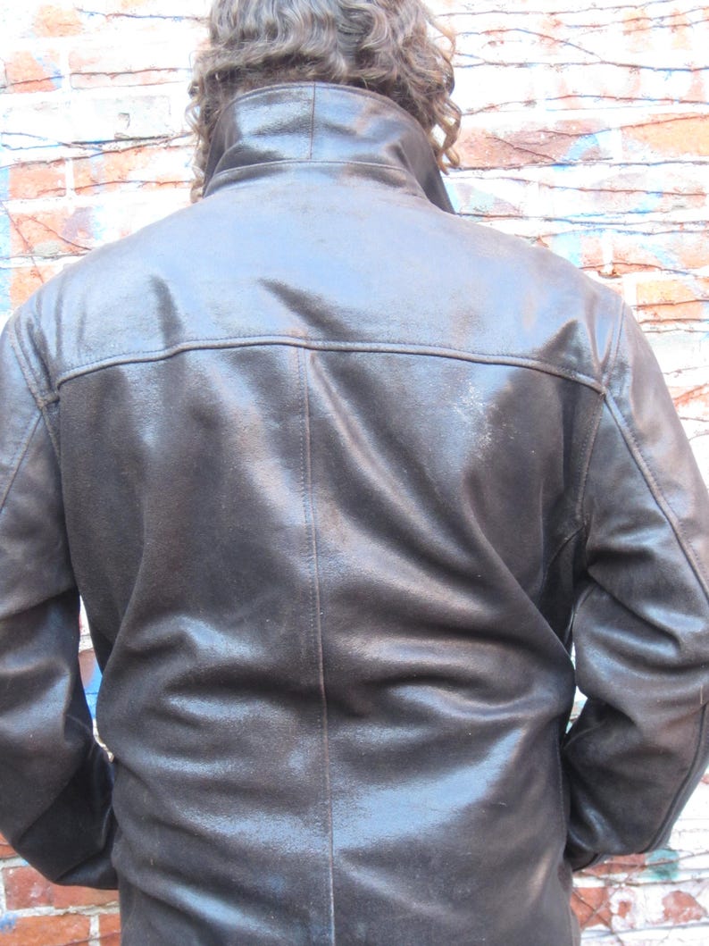 May include: A dark brown leather jacket with a high collar and a glossy finish. The jacket has a classic design with visible stitching and a tailored fit. The image shows the back of the jacket, highlighting its structured shoulders and sleek silhouette.