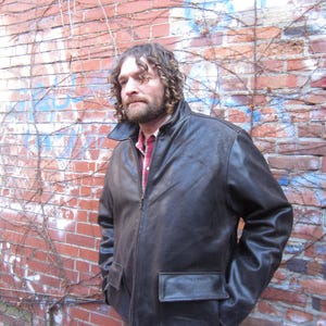 May include: A dark brown leather jacket with a zipper closure and two front pockets. The jacket has a classic design and a collared neckline. The person is wearing a red plaid shirt underneath. The background is a brick wall with vines.
