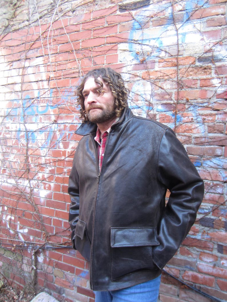 May include: A dark brown leather jacket with a zipper closure and two front pockets. The jacket has a classic design and a collared neckline. The person is wearing a red plaid shirt underneath. The background is a brick wall with vines.
