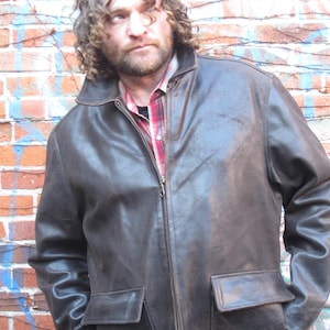 May include: A dark brown leather jacket with a zip-up front and two front pockets. The jacket has a collared neckline and long sleeves. The person is wearing a red and black plaid shirt underneath. The background is a brick wall with some blue and white paint.