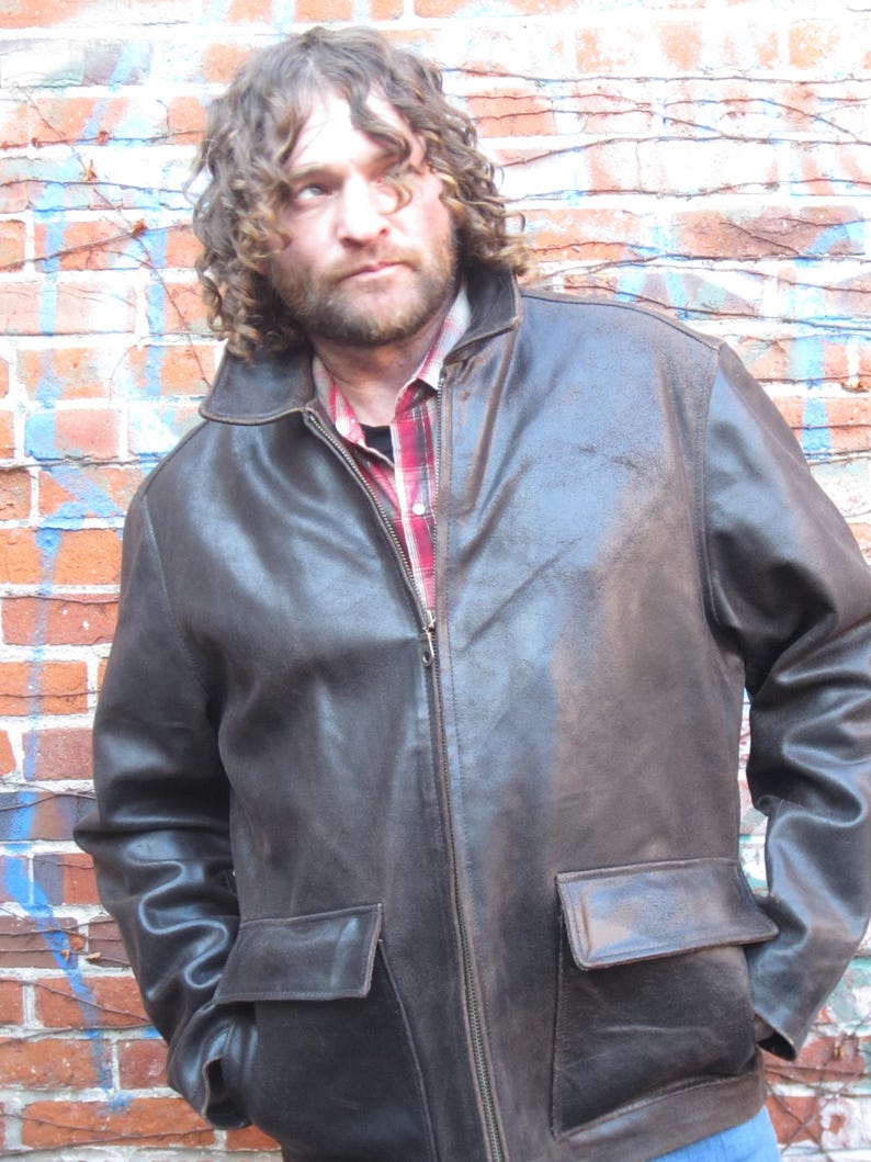 May include: A dark brown leather jacket with a zip-up front and two front pockets. The jacket has a collared neckline and long sleeves. The person is wearing a red and black plaid shirt underneath. The background is a brick wall with some blue and white paint.