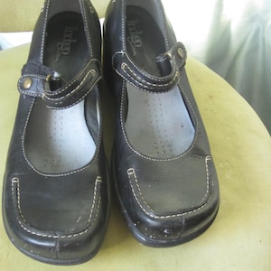 May include: A pair of black leather Mary Jane style shoes with white stitching and a strap closure. The shoes have a rounded toe and a small heel. The brand name "indigo" is visible on the insole.