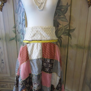 Op de afbeelding: Een patchwork rok met een gele tailleband. De rok is gemaakt van verschillende stoffen in tinten roze, rood, bruin en wit. De rok is op een witte mannequin.