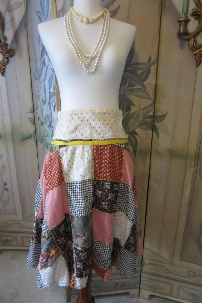 Op de afbeelding: Een patchwork rok met een gele tailleband. De rok is gemaakt van verschillende stoffen in tinten roze, rood, bruin en wit. De rok is op een witte mannequin.