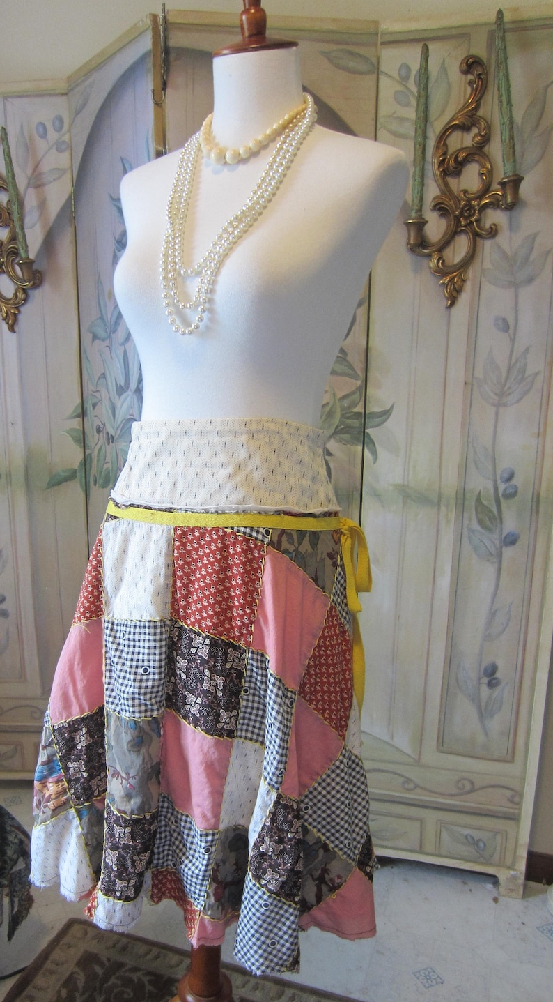 Op de afbeelding: Een witte paspop met een gele en witte patchworkrok. De rok is gemaakt van verschillende stoffen en heeft een gele band die om de taille is geknoopt. De paspop draagt een drievoudige witte parelketting.