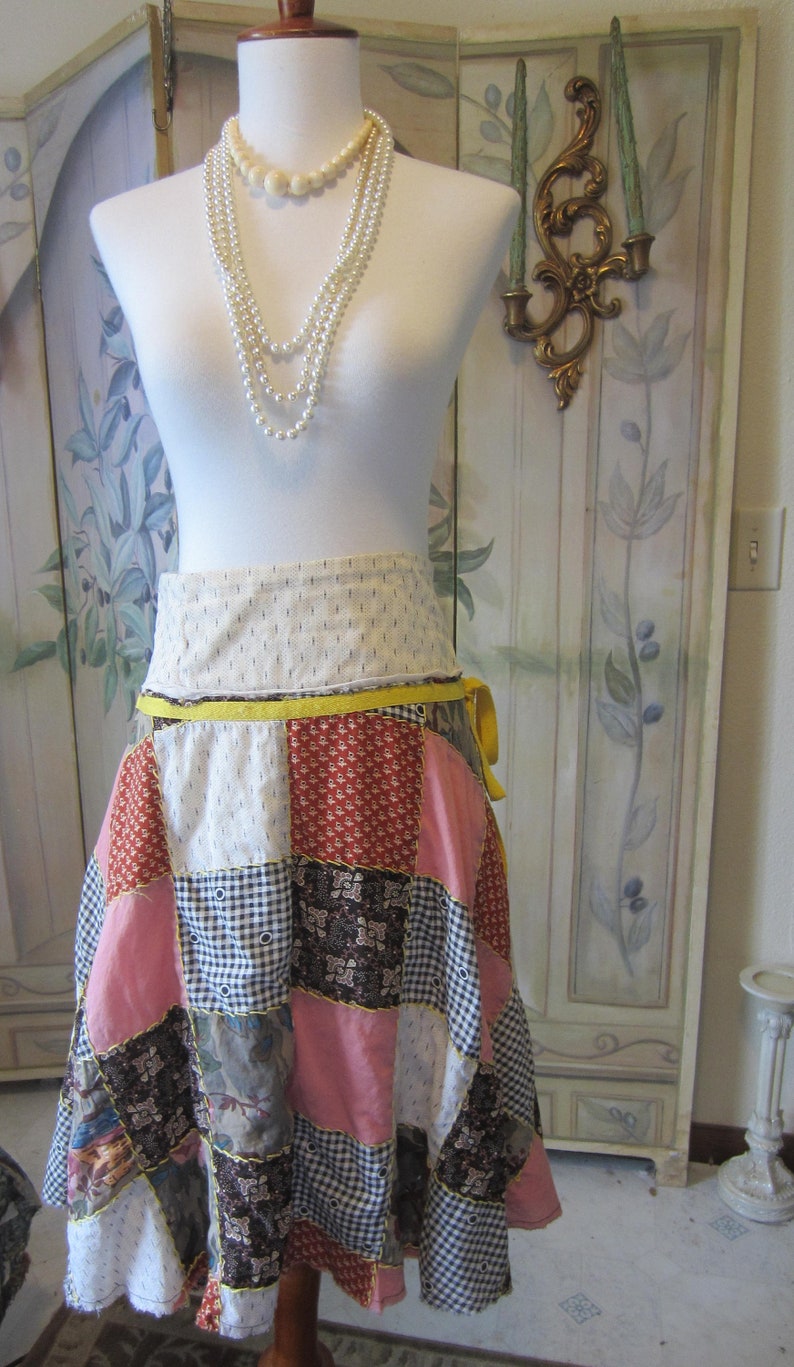 Op de afbeelding: Een witte paspop met een veelkleurige patchwork rok. De rok heeft een gele tailleband en een gele strik. De paspop draagt een meerrijige parelketting.