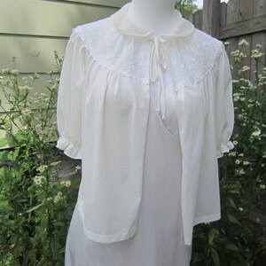 Pode incluir: Um camis&atilde;o branco longo e sedoso com uma bata branca combinando com acabamento em renda. A bata tem um la&ccedil;o no pesco&ccedil;o.