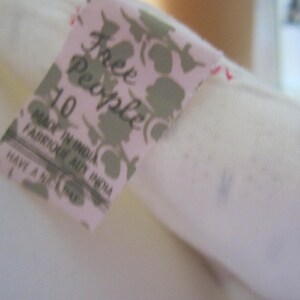 Op de afbeelding: Een wit Free People kledinglabel met een bloemenpatroon en de tekst "Free People" en "Made in India" in zwart. Het label bevat ook de maat "10" en de tekst "Have a nice day".