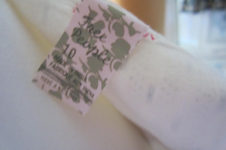Op de afbeelding: Een wit Free People kledinglabel met een bloemenpatroon en de tekst "Free People" en "Made in India" in zwart. Het label bevat ook de maat "10" en de tekst "Have a nice day".