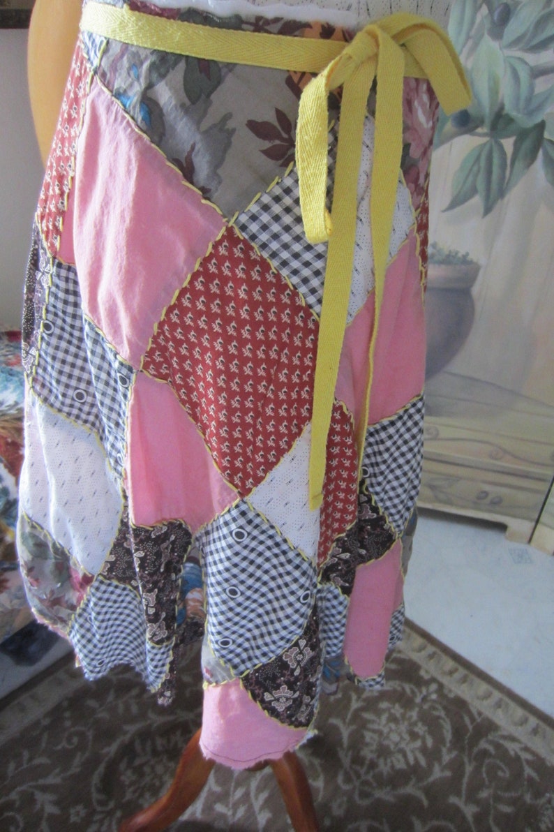 Op de afbeelding: Een patchwork wikkelrok met een gele stoffen strik. De rok is gemaakt van verschillende stoffen in roze, rood, wit en zwart.