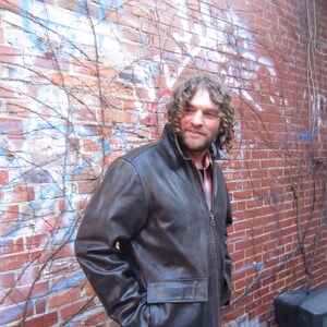 May include: A brown leather jacket with a zip-up front and two front pockets. The jacket has a collar and is worn over a red and black plaid shirt. The background is a red brick wall with some graffiti and vines.