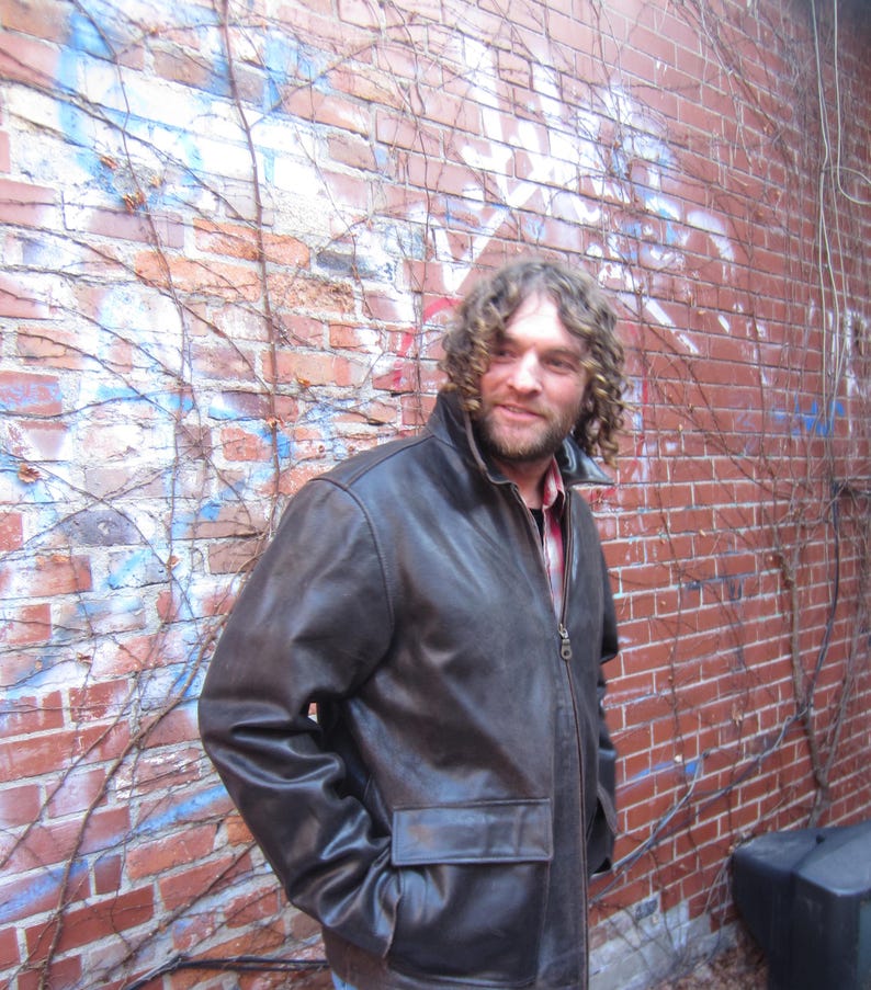 May include: A brown leather jacket with a zip-up front and two front pockets. The jacket has a collar and is worn over a red and black plaid shirt. The background is a red brick wall with some graffiti and vines.