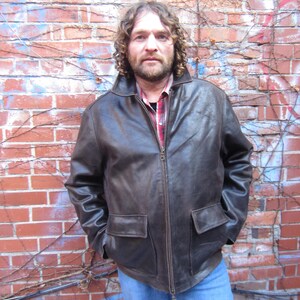 May include: A brown leather jacket with a zip-up front and two front pockets. The jacket has a collared neckline and is worn over a red and black plaid shirt and blue jeans. The background is a brick wall with vines.