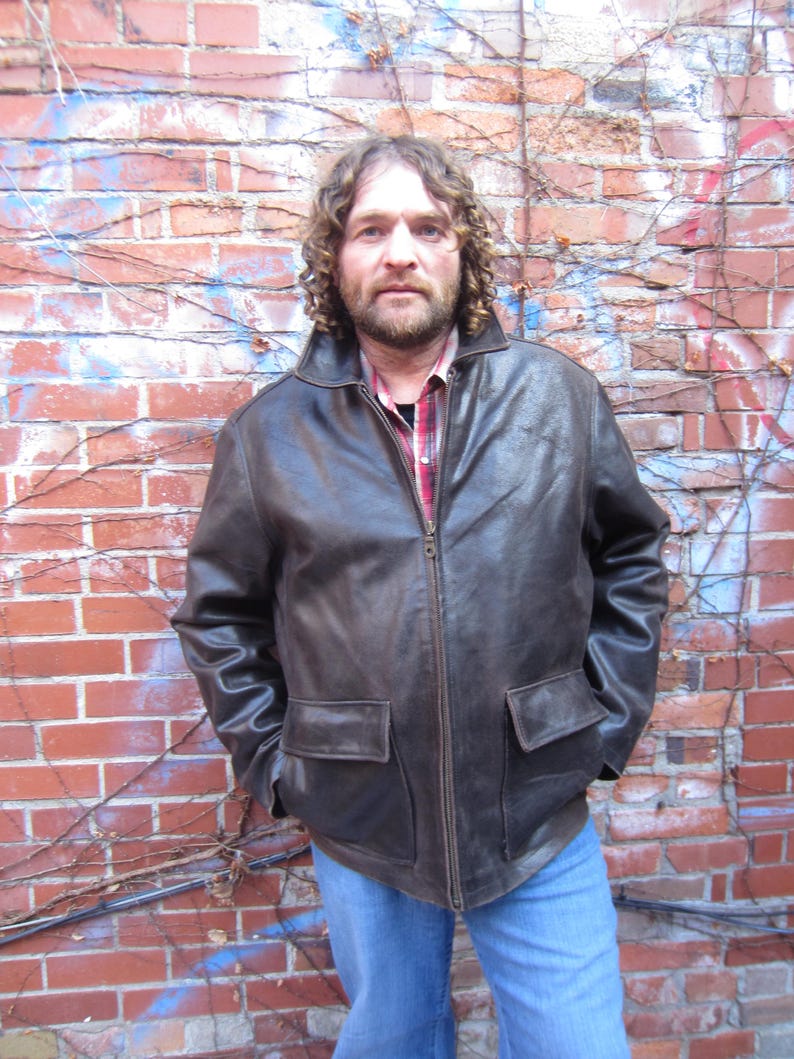 May include: A brown leather jacket with a zip-up front and two front pockets. The jacket has a collared neckline and is worn over a red and black plaid shirt and blue jeans. The background is a brick wall with vines.