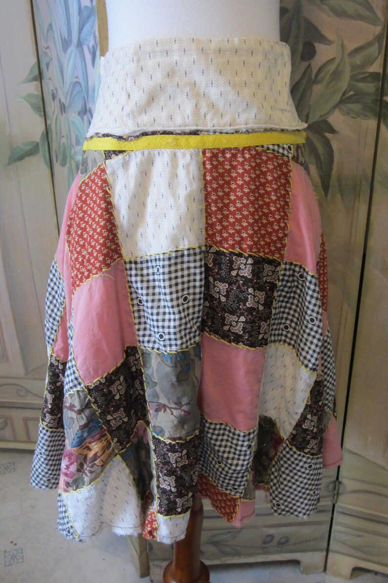 Op de afbeelding: Een patchwork rok met verschillende bloemen- en geometrische patronen in tinten roze, rood, bruin en wit. De rok heeft een gele tailleband en is gemaakt van katoenen stof.