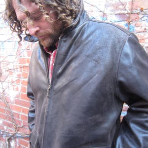 May include: A dark brown leather jacket with a zipper closure and two front pockets. The jacket has a collar and is worn over a red and white plaid shirt. The person wearing the jacket is standing in front of a brick wall.
